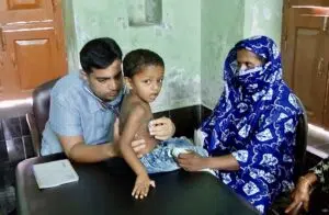 Dr. Md. Abdullah Yousuf performing a pediatric clinical evaluation with a stethoscope on a young child accompanied by his mother during a primary care health assessment.