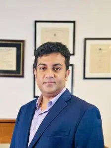 Dr. Md. Abdullah Yousuf, Chief Medical Consultant at Back in Motion, wearing a blue blazer and light purple shirt, posing in front of framed medical credentials in Dhaka.