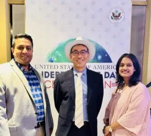 Dr. Md. Abdullah Yousuf and Dr. Tazia Sardar standing with an international diplomat in front of a United States of America Independence Day Celebration banner in Dhaka.